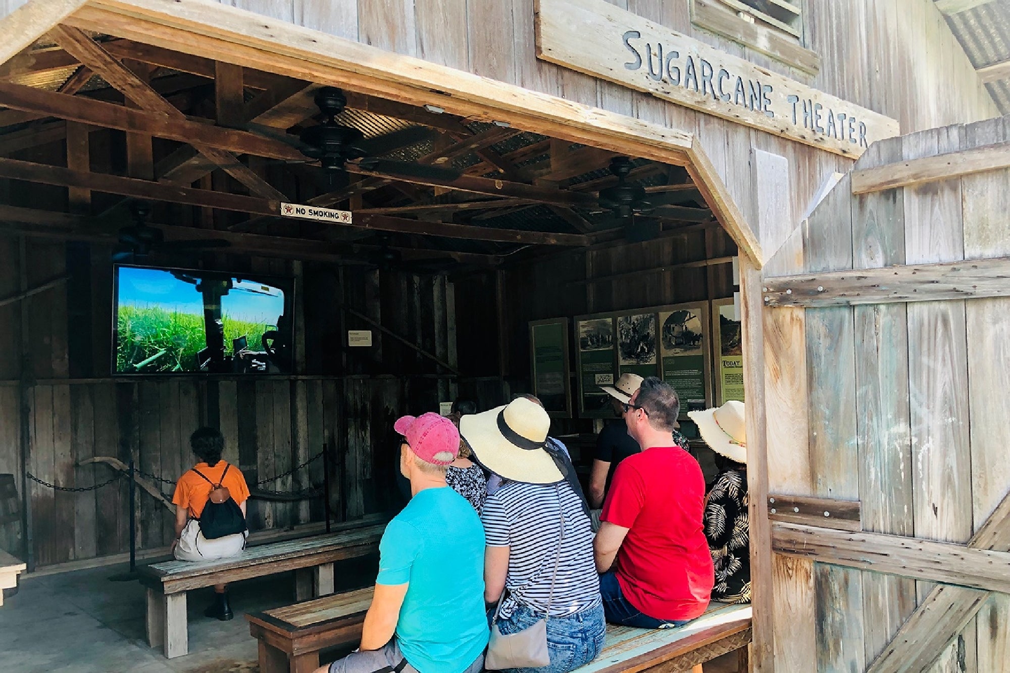 A group of people sit on benches in a wooden barn labeled "Sugarcane Theater," watching a video on a screen at the front.