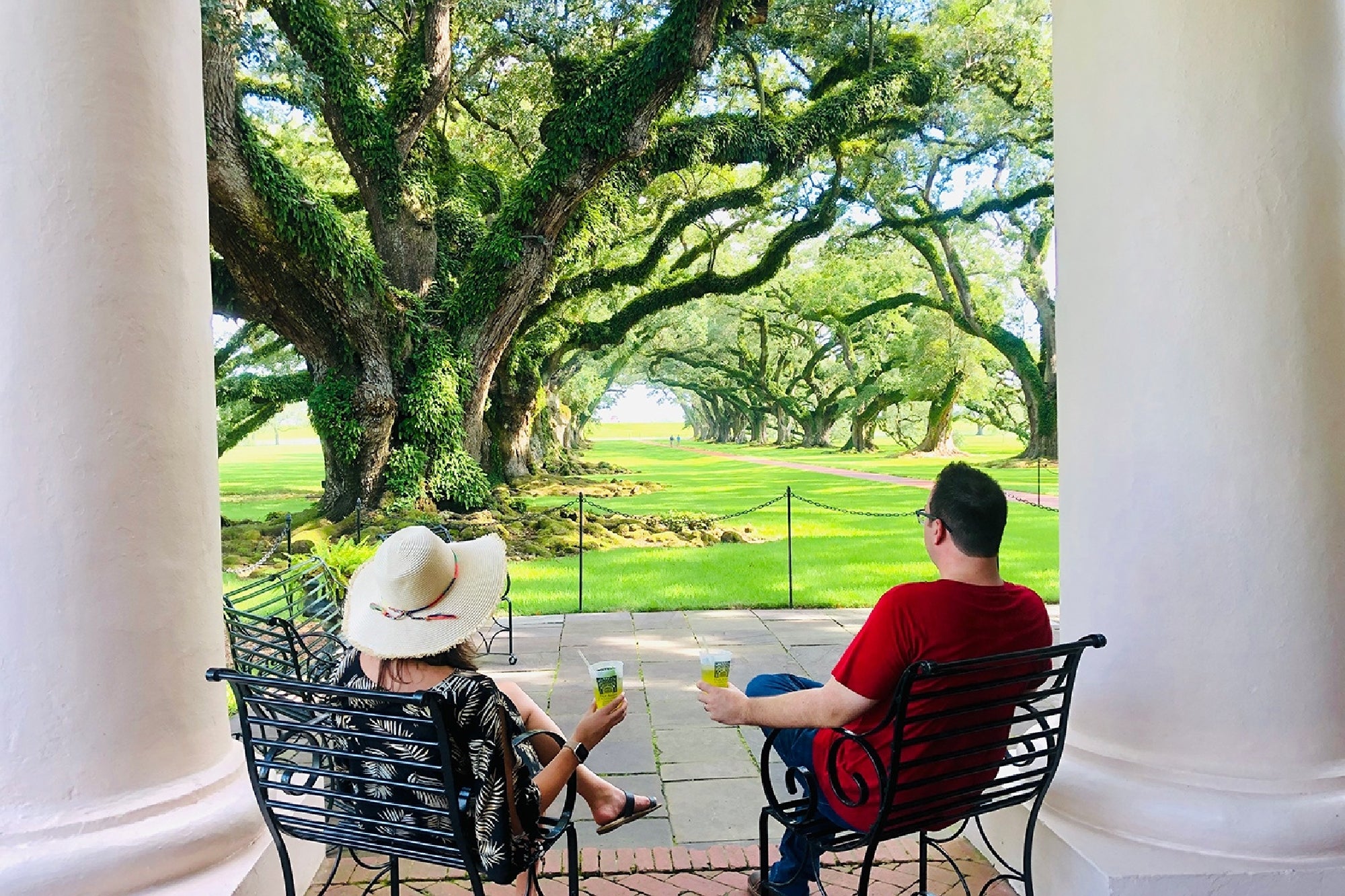 Two people sit on wrought iron chairs with drinks, facing a lawn lined with large, sprawling oak trees, framed by white columns.