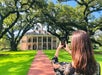 A woman stands on a brick pathway taking a photo of a large, historic plantation house with columns and balconies, surrounded by oak trees and green lawn.