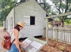 A woman wearing a hat and backpack reads an informational sign in front of a historic wooden building surrounded by trees and a picket fence.