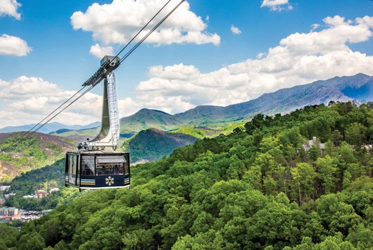 The Ober Mountain Tram suspended above green forests with the Smoky Mountains and a blue sky with fluffy clouds in the background.