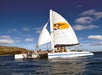 A white catamaran with people on board sails on the ocean near a rocky coastline under a blue sky. The sail features an illustration of a woman in a yellow circle.