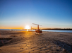 A helicopter is parked on a sandy beach at sunset, with the sun setting over the ocean horizon in the background.