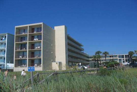 Four-story beachfront hotel with balconies, surrounded by grass and parked cars. Clear blue sky in the background. Room balconies overlook a sandy area.