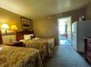 Hotel room with two double beds, patterned bedspreads, a nightstand with lamps, a painting above, a refrigerator, a dresser, and a view of the bathroom and windowed area in the background.