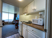Small kitchenette with a stove, sink, microwave, and coffee maker. Cabinets above and below the counter. Living area visible with seating, table, and a TV near a large window.