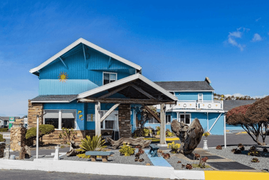 The Oceanside Inn & Suites exterior with blue siding with white accents and lots of landscaping on a sunny day.