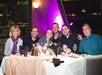 Six people sit together at a restaurant table with drinks, napkins, and utensils, smiling at the camera under dim, purple lighting.