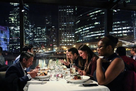 People dining at an elegant restaurant with a city skyline visible through large windows in the background.
