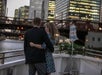 A couple stands arm-in-arm on a boat, looking at a city skyline with lit-up skyscrapers and a bridge over a river.
