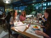 A group of people sitting at a restaurant table. A woman smiles while holding a dessert with "Happy Birthday" written on it. Others are engaging and taking photos.