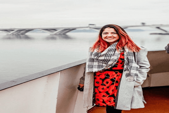 Smiling person with red hair stands on a boat wearing a grey coat and patterned scarf. A bridge is visible in the background over water.