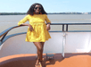 Person in a yellow dress stands on a boat deck by the water, wearing sunglasses and smiling.
