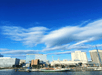 A city skyline with modern buildings under a blue sky and scattered clouds. A boat is docked in the foreground near a waterfront.
