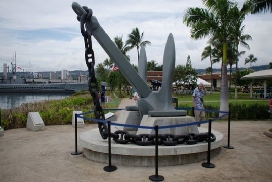 Large gray ship anchor displayed outdoors on a concrete platform, surrounded by a chain, palm trees, and a roped barrier; people and buildings in the background.