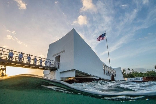 White memorial structure with an American flag, partly above water, with people standing on a bridge at sunset; the USS Arizona Memorial at Pearl Harbor.