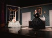 Two people in period costumes perform on a stage set designed as an elegant 19th-century room, featuring patterned walls, a desk, a sofa, and a portrait above a fireplace.