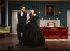 Two actors in period costumes perform in a room with 19th-century decor, a portrait of George Washington, and a wooden desk. The man looks discontented while the woman gestures toward him.