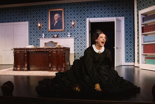 A woman in a long black dress kneels on a stage floor, screaming, in an ornate office set with a large desk, open door, and a portrait of George Washington on the wall.