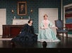 Two actors in period costumes perform on stage. One sits in a striped dress on a chair, the other kneels in a dark dress. A portrait and a desk are in the background.