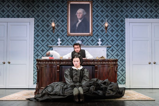 A woman in a black dress sits on the floor in front of a large wooden desk, with two men behind it and a portrait of George Washington on the wall.