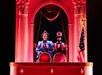 Two people in period costumes stand in a theater box with red lighting, columns, and a U.S. flag nearby.