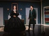 Two actors in period costumes perform on stage; one stands with a hand on their chest in a long black dress, while the other, in a green coat, looks on. Bookshelves and a fireplace are in the background.