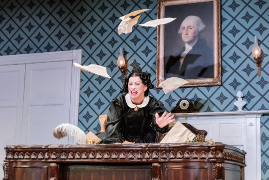 A woman in period costume sits at a large wooden desk, throwing papers into the air, with a portrait of George Washington hanging on the patterned wall behind her.
