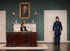 A man in period attire sits at a wooden desk in a vintage office setting with a painting of George Washington. Another man in a blue uniform stands near a door against a patterned wallpaper.