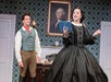 A woman in a black Victorian dress reads from a book while a man in historical attire stands behind her; a portrait hangs on the wall in a patterned room.