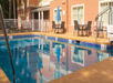 Outdoor pool area with tables and chairs around the deck. Nearby, there is a building with large windows and an accessible pool lift.