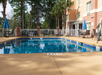 Outdoor pool area with several tables and chairs, surrounded by trees, with a sign on the fence.