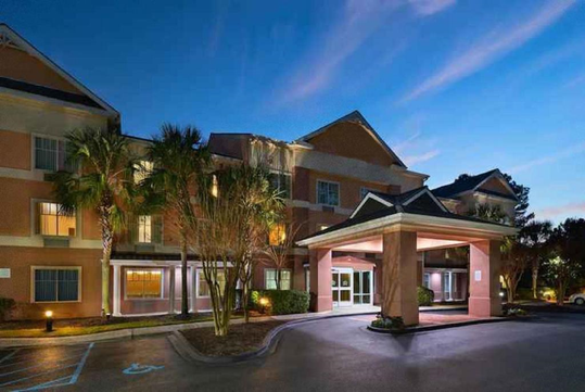 A three-story hotel with a covered entrance, lighting on, surrounded by palm trees at dusk.