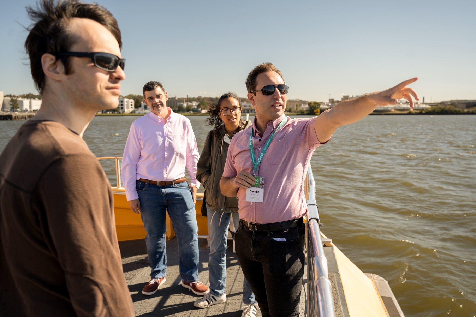 Four people stand on a boat deck by the water; one man in sunglasses and a pink shirt points toward something in the distance.