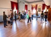 People stand in pairs facing each other in a spacious, sunlit room with wooden floors, red curtains, chandeliers, and large windows, participating in an activity or class.