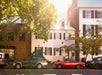 Two cars parked on a sunny street in front of a white townhouse with green shutters and an American flag; trees cast shadows on the buildings and sidewalk.
