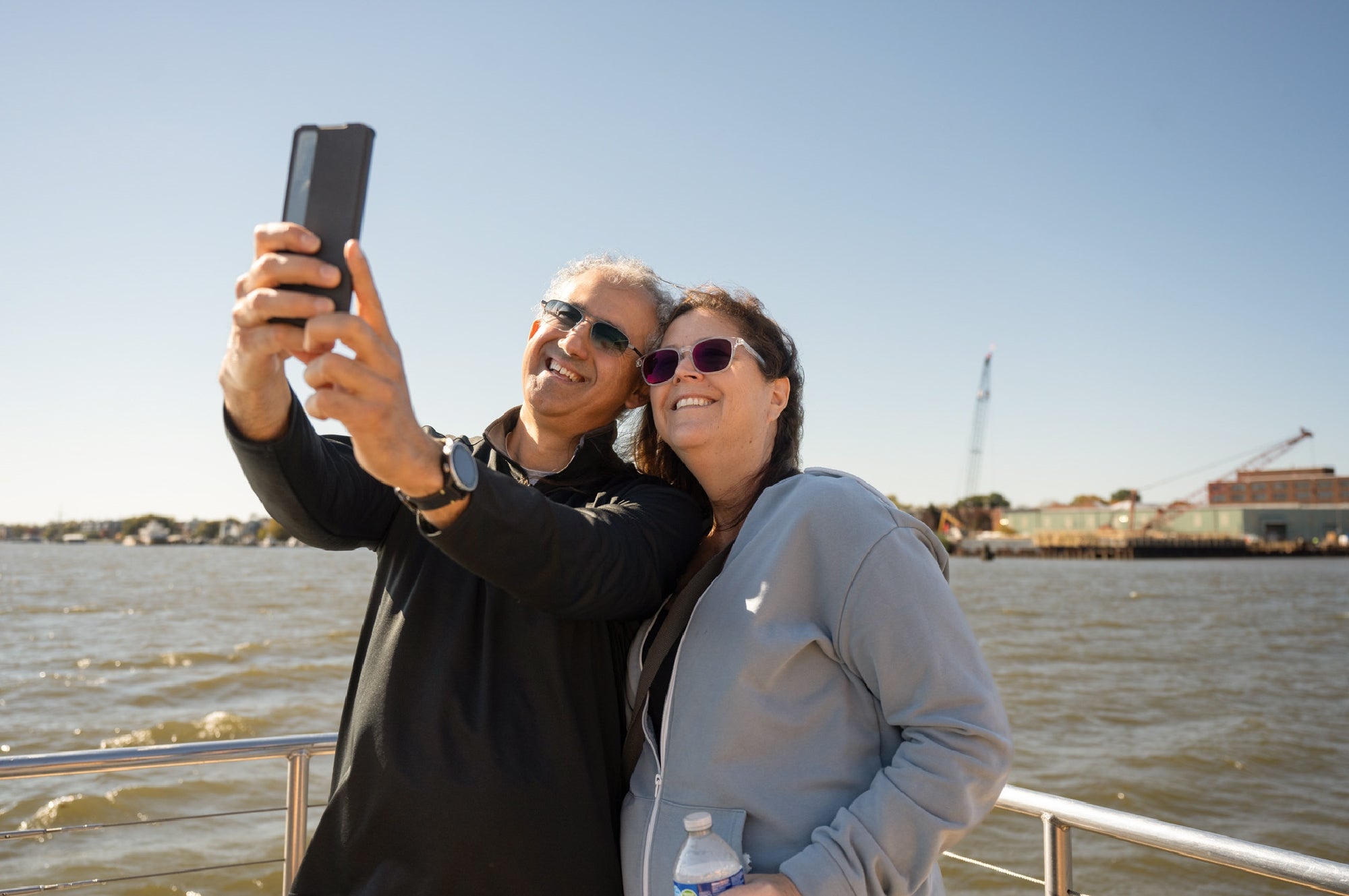 Two people wearing sunglasses take a selfie together on a boat, with water, industrial buildings, and a crane visible in the background under a clear sky.
