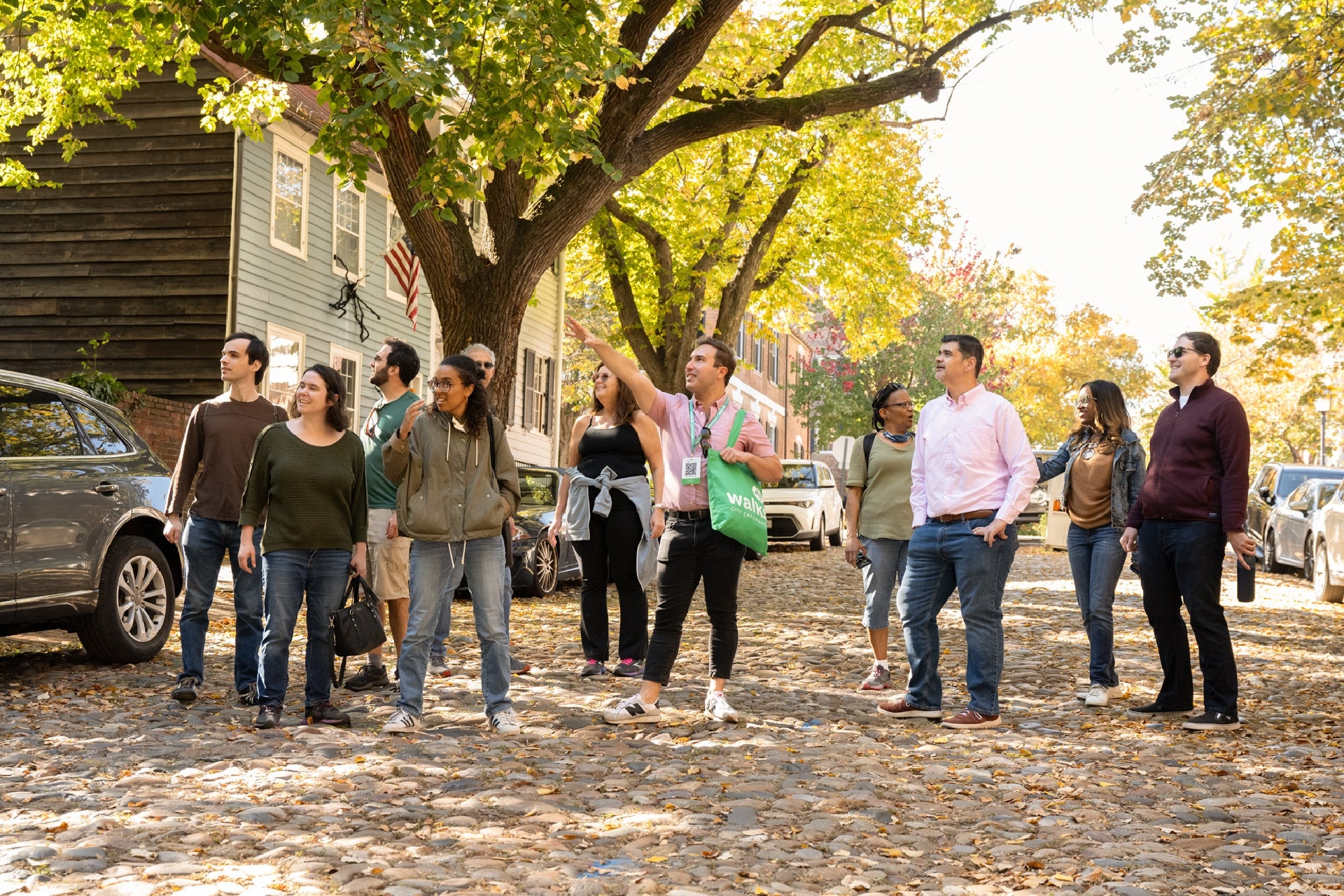 A group of people stands on a cobblestone street under trees, with one person in the center pointing upward, as others look around or ahead. Cars and houses line the background.