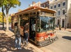 Several people board a red and gold trolley bus stopped at a curb on a sunny day in a historic city neighborhood.