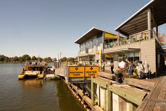 A modern waterfront building with people gathered outside, next to a dock with yellow sightseeing tour and water taxi boats on a sunny day.