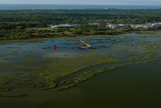 A colorful helicopter flies over a marshland with scattered greenery, with dense forest and buildings visible in the background.