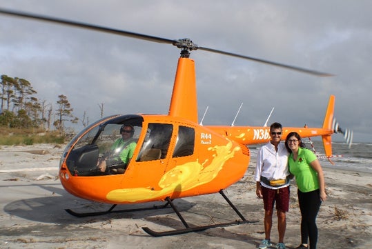 Two people stand in front of an orange helicopter on a sandy area with another person visible inside the cockpit; trees and cloudy sky in the background.
