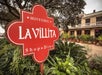 Sign for "La Villita" in a historic district with greenery in the foreground and a building with potted plants in the background.