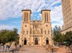 Facade of a historic church with twin towers, a rose window, and a plaza with tables and trees in the foreground.