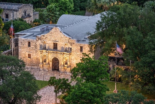 Aerial view of an old stone building with an arched entryway and flagpoles, surrounded by trees and greenery.