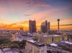 A cityscape at sunset featuring a skyline with modern buildings and a distinctive tower on the right, under a colorful sky with orange and purple hues.