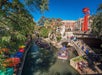 Boats with tourists navigate the river surrounded by colorful umbrellas near a stone bridge and buildings under a clear blue sky.