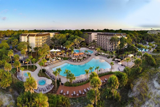 Aerial view of a resort with two large pools, surrounded by lounge chairs and greenery, with several buildings in the background under a clear sky.
