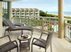Balcony view with wicker chairs and loungers, a table with wine and snacks, overlooking a pool, greenery, and the ocean in the background.