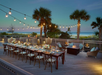 An outdoor dining area on a deck overlooks the ocean at sunset. A long table is set with flowers and place settings. String lights and palm trees add to the ambiance.